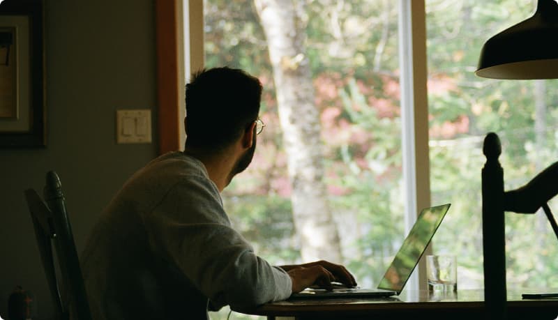 Mies istuu pöydän ääressä tietokoneella ja katsoo ulos ikkunasta.