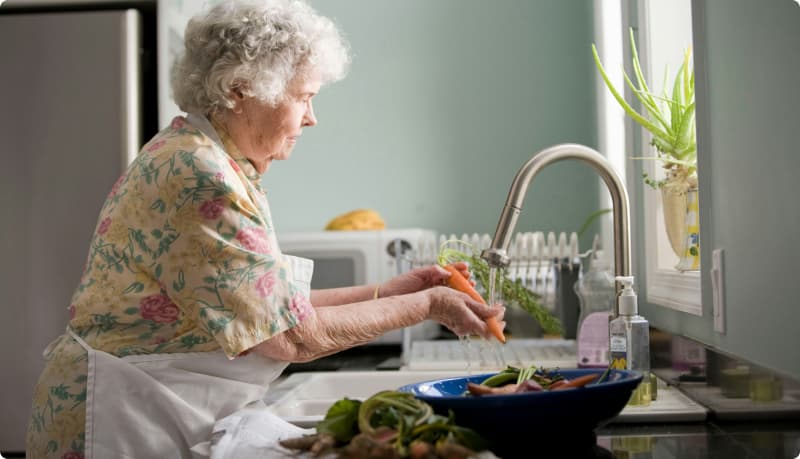 Harmaanhiuksinen nainen laittaa ruokaa keittiössä ja pesee vihanneksia.