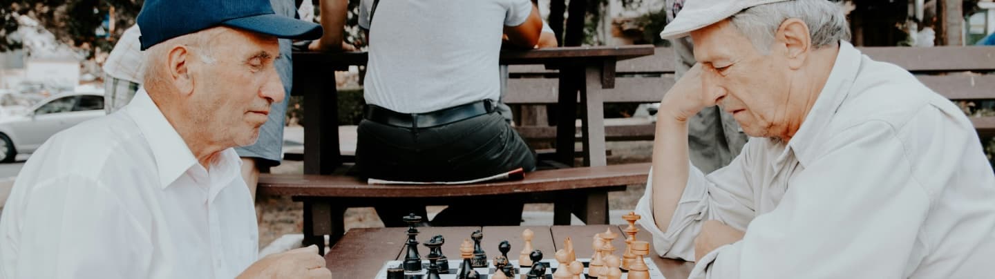 Kaksi vanhempaa miestä pelaa ulkona puistossa shakkia.