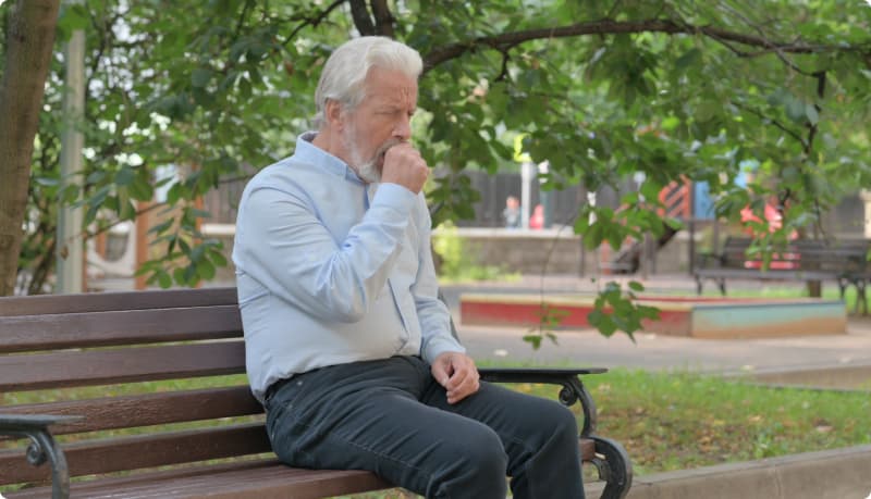 Mies istuu puiston penkillä ja pitää kättään suun edessä yskien.