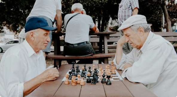 Kaksi vanhempaa miestä pelaa ulkona puistossa shakkia.